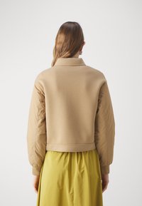 Brązowa kurtka bomberka z fakturowanej, gładkiej tkaniny, z żebrowanymi mankietami i krótkim dołem, w zestawieniu z zwiewną żółtą spódnicą.