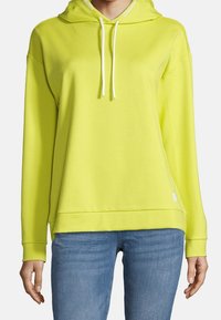 Person som bär en neongul huvtröja med vita snoddar och blå jeans, står med armarna avslappnat längs sidorna.