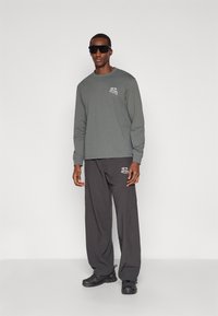 Grijze longsleeve met grafische print aan de voorkant, gecombineerd met loszittende donkere broek en zwarte sneakers. Model draagt een zwarte zonnebril.