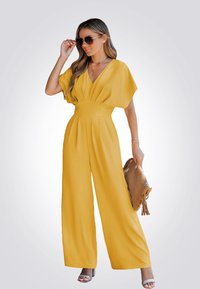 Combinaison ample jaune avec un décolleté en V profond, des manches courtes et un tissu texturé. Les accessoires comprennent une pochette marron et des lunettes de soleil.