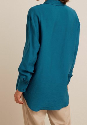 Chemise turquoise à manches longues avec col, tissu légèrement texturé, et poignets boutonnés, portée avec un pantalon beige. Vue de dos.