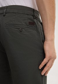 Dunkelgraue Hose aus einem leichten Stoff mit glatter Textur, ausgestattet mit einer Gesäßtasche mit Knopf und einem markierten Lederpatch.