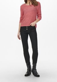 Rosa Strickpullover mit rundem Ausschnitt, kombiniert mit schwarzen Skinny-Jeans mit Knopfleiste vorne. Schwarze Stiefeletten vervollständigen das Outfit.