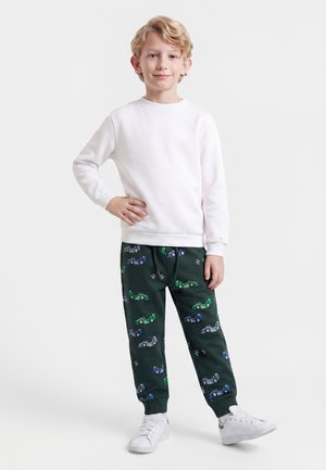 Enfant blond debout, main sur la hanche, portant une chemise blanche à manches longues, pantalon foncé avec des imprimés de voitures, et des baskets blanches sur fond uni.
