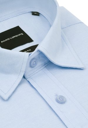 Jasnoniebieska bawełniana koszula z klasycznym kołnierzykiem, pojedynczym zapięciem na guzik oraz czarnymi metkami z logo wskazującymi krój i projektanta.