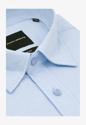 Jasnoniebieska bawełniana koszula z klasycznym kołnierzykiem, pojedynczym zapięciem na guzik oraz czarnymi metkami z logo wskazującymi krój i projektanta.