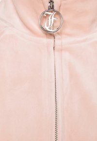 Gros plan d'un vêtement rose pâle avec une fermeture éclair argentée et un tire-zip rond argenté orné d'une lettre F décorative.