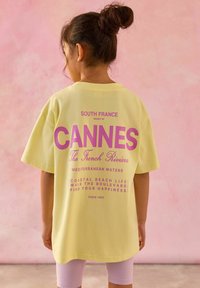 Žuta pamučna majica s ružičastim natpisima na leđima, uključujući "CANNES" i druge fraze, u kombinaciji s svijetloplavim šortsem.