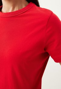 Nærbillede af en person iført en ensfarvet rød t-shirt med korte ærmer og rund hals mod en lys baggrund.