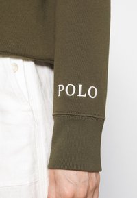 Olivgrön långärmad skjorta med ribbad mudd och vit broderad "POLO" text på ärmen. Slät bomullstyg.