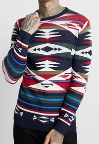 Sweter dzianinowy z okrągłym dekoltem, z wielokolorowym wzorem geometrycznym w kolorach granatowym, czerwonym, zielonym i białym, wykonany z miękkiego materiału.