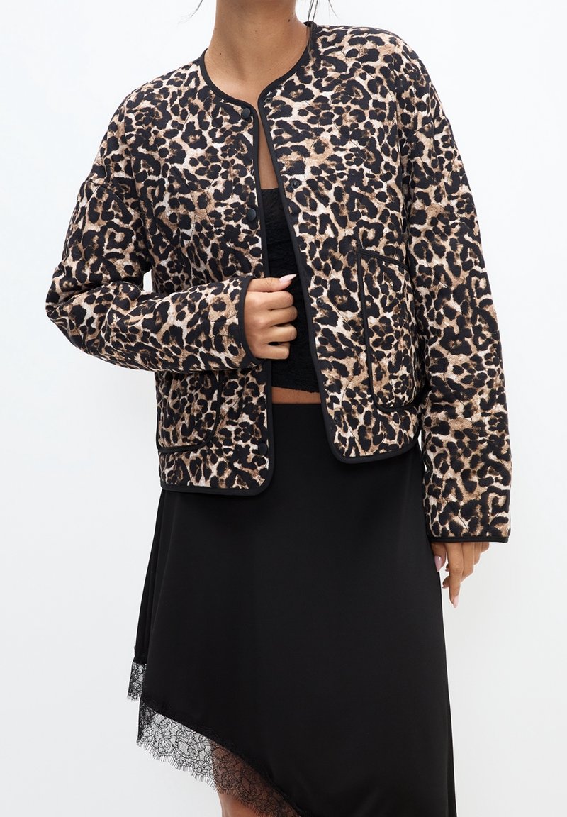 Femme portant une veste boutonnée à imprimé léopard sur une jupe noire bordée de dentelle et un haut noir, sur fond blanc.