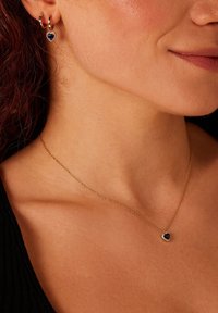 Goldene Kettenhalskette mit einem herzförmigen blauen Anhänger und einem schimmernden Halo. Passende goldene Creolen mit kleinen blauen Edelsteinen.