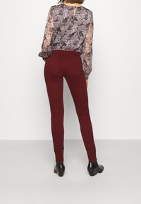 Jeans skinny burdeos de cintura alta, con costuras en contraste y dos bolsillos traseros, combinados con una blusa de patrón semitransparente.