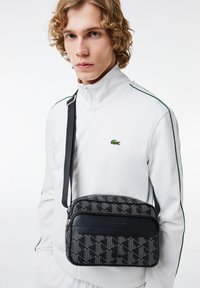 Czarna torebka crossbody w geometryczny wzór z teksturowanymi skórzanymi akcentami, z zamkiem błyskawicznym na górze i regulowanym paskiem. Modelka nosi białą odzież.