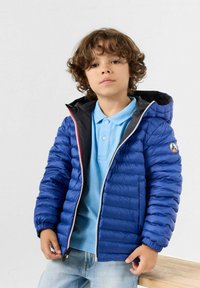 Giacca a vento blu con cappuccio, dotata di chiusura con zip, foderatura nera a contrasto e un emblema sulla manica sinistra. Indossata sopra una polo celeste.