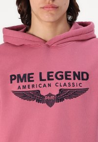 Różowa bluza z kapturem, z sznurkiem, z ciemnonawigacyjnym haftowanym logo "PME LEGEND AMERICAN CLASSIC" oraz grafikami skrzydeł z przodu.
