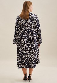Robe longue navy avec un motif abstrait beige, manches longues et taille nouée. Tissu lisse, longueur cheville, assortie à des talons noirs.