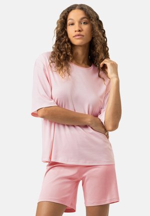 Junge Frau mit lockigem Haar, die ein hellrosa gestreiftes Kurzarmoberteil und passende rosa Shorts trägt, steht mit einer Hand nahe ihrem Gesicht.