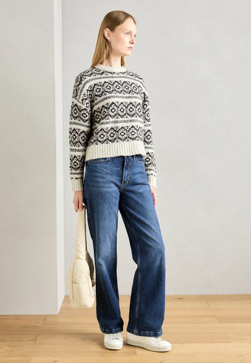 Strickpullover in Creme mit schwarzem geometrischem Muster, kombiniert mit weiten blauen Jeans und weißen Sneakers, hält eine cremefarbene Umhängetasche.