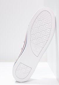 Biała podeszwa tenisówki Converse All Star z geometrycznym wzorem bieżnika, z czerwonymi i niebieskimi pasami po bokach, ustawiona pionowo na białym tle.