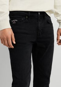 Osoba oblečená v čiernych Pepe Jeans London rifliach a bielej košeli s dlhým rukávom, ruky uvoľnene pozdĺž strán.
