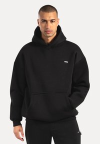 Zwarte capuchontrui van zachte stof, met een borstzak aan de voorkant, geribbelde manchetten en een klein logoplaatje op de borst.