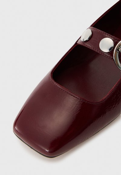 Burgundy patent leather shoe with a square toe and open sides. Features a strap with silver hardware accents and a circular buckle.