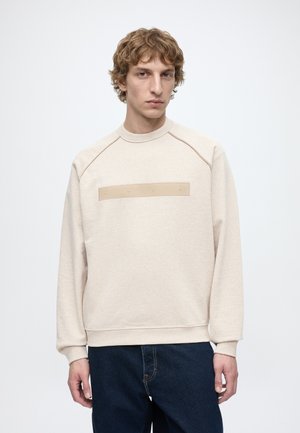 Young man with light curly hair wearing a beige sweatshirt with a rectangular patch and dark blue jeans, standing against a plain background.