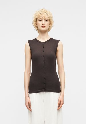 Blonde Frau mit kurzen lockigen Haaren, die ein ärmelloses schwarzes, geknöpftes Oberteil und lockere weiße Hosen trägt, vor einem einfarbigen Hintergrund stehend.