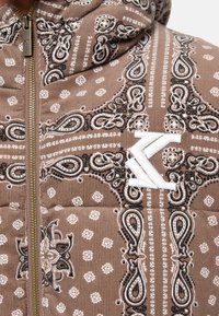 Brązowa kurtka ocieplana z wzorem paisley i geometrycznym, złotym zamkiem błyskawicznym oraz wyraźnym, białym haftowanym logo z przodu.