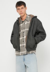 Sweatshirt à capuche gris foncé avec une doublure beige, porté par-dessus une chemise à carreaux dans des tons blancs, bruns et bleus. Coupe décontractée.