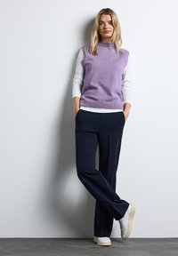 Lila ärmlös knittröja över en vit långärmad skjorta, kombinerad med marinblå vida byxor och vita sneakers. Släta, lager på lager texturer.