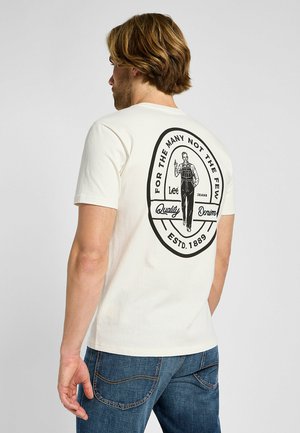 Kremowa bawełniana koszulka z czarnym graficznym wzorem na plecach, przedstawiająca postać i napis: „DLA WIELU, NIE DLA NIELICZNYCH, Lee Jeans, Jakość Dżinsu, ZAŁOŻONA 1889.”