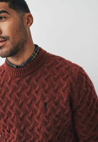Maglione a maglia color bordeaux con un motivo a diamante testurizzato, caratterizzato da un collo a girocollo a costine e un piccolo logo sul petto, indossato sopra una camicia a quadri.