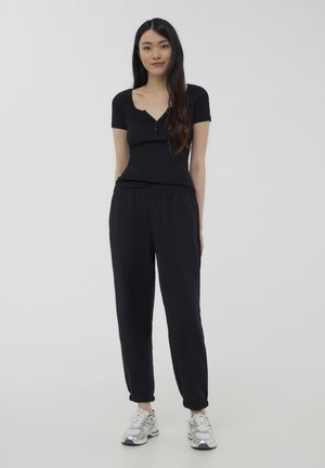 Body corto nero a maniche corte con scollo a V e dettagli con bottoni, abbinato a pantaloni jogger neri con vita elasticizzata. Il modello indossa sneakers argentate.