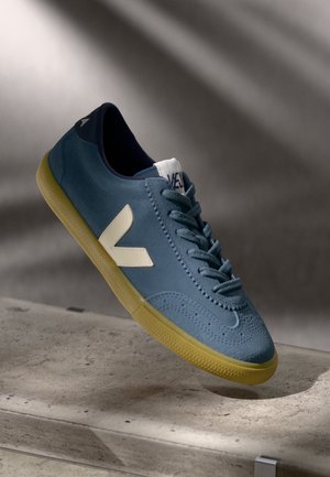 Blue suede sneaker with cream logo, textured upper, padded collar, and yellow rubber sole. Features classic lacing and perforated accents.