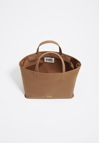 Bolso tote de tela color tostado con dos asas superiores, que cuenta con un forro interior y un acento de logo dorado en la parte delantera. Diseño abierto, sin cierre visible.