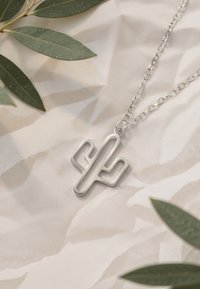 Ciondolo d'argento a forma di cactus su una catena, con un design minimalista e linee morbide, poggiante su uno sfondo beige chiaro con foglie.