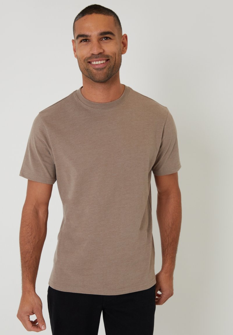 Hombre sonriente con cabello corto, vestido con una camiseta marrón lisa de manga corta y pantalones negros, sobre un fondo gris claro.