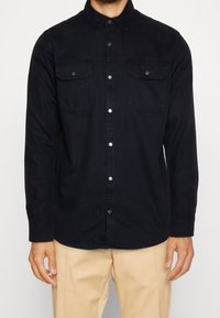 Chemise noire à boutons, à manches longues, avec deux poches poitrine, boutons pression et texture douce, associée à un pantalon beige.