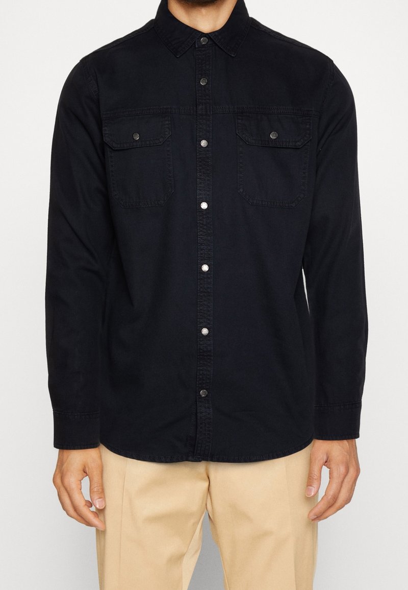 Chemise noire à boutons, à manches longues, avec deux poches poitrine, boutons pression et texture douce, associée à un pantalon beige.
