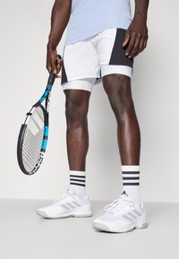 Pantalones cortos deportivos blancos y negros, camiseta corta azul, zapatillas blancas con detalles grises, calcetines a rayas, sosteniendo una raqueta de tenis.