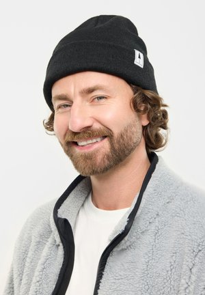 Schwarze Strickmütze mit einem kleinen weißen Etikett, das ein Baumdesign zeigt. Die Person trägt eine graue Fleecejacke mit einem schwarzen Reißverschluss und einem weißen Shirt darunter.