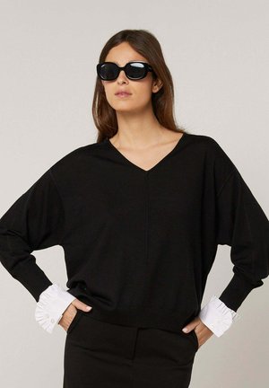Femme portant un pull noir à col en V avec des poignets blancs froufroutants, une jupe noire et de grandes lunettes de soleil noires, posant avec les mains dans les poches.