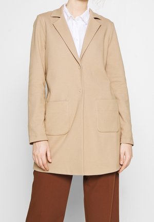 Blazer beige fabriqué en tissu texturé, avec un col à revers, deux grandes poches avant et une fermeture à un bouton.