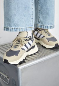 Sneaker beige e grigio con strisce bianche, caratterizzata da un mix di materiali scamosciati e mesh, suola spessa e lacci rotondi.