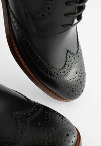 Zapatos brogue de cuero negro con detalles perforados, bordes curvados y suelas marrones. Los cordones planos son visibles en la parte central superior.