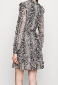 Frau trägt ein tailliertes, langärmeliges, knielanges Kleid mit grauem und schwarzem Schlangenprint und Rüschensaum, von hinten gezeigt.