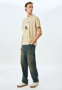 Beige kortærmet t-shirt med et trykt design, der viser en jordbær og en lime. Bærers sammen med løstsiddende blå denimbukser og hvide sneakers.